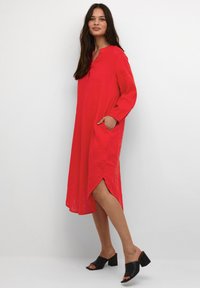 Röd långärmad klänning med V-ringning, sidofickor och slitsar i fållarna. Slät textur. Bärs med svarta öppna muleklackar.