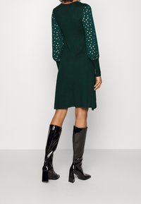 Groene gebreide jurk met lange, puffed mouwen, versierd met metallic gouden stippen. Gecombineerd met zwarte, glanzende kniehoge laarzen en een getailleerde pasvorm.