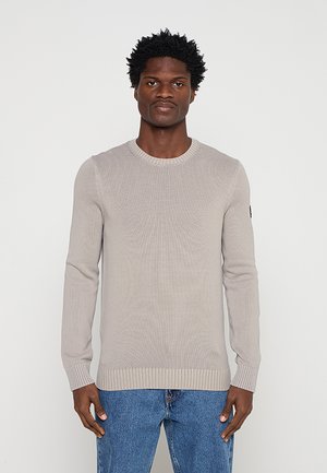 Hombre con un suéter de canalé beige claro y jeans azules, de pie frente a un fondo blanco liso.