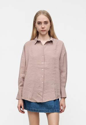 Jeune femme aux longs cheveux blonds porte une chemise rose ample boutonnée et une courte jupe en jean bleue, debout devant un fond blanc simple.