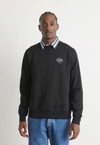 Fekete, pamut keverékből készült pulóver, kerek nyakkal, bordás mandzsettákkal, a "Tommy Hilfiger" logóval a mellkason, csíkos ingre rétegzett.