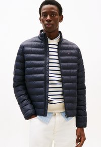 Jeune homme portant une veste matelassée bleu marine par-dessus un pull rayé et une chemise bleu clair, avec un pantalon blanc, debout devant un fond blanc uni.