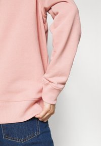 Sudadera rosa claro con puños y dobladillo acanalados, con un material de punto texturizado. Una mano agarra la tela en la cintura, combinada con jeans azules.