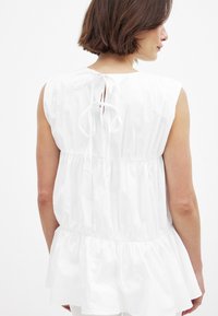 Valge ilma varrukateta pluus, millel on kogutud talje, peplum ääre ja taga sõlme detail. Valmistatud kergest puuvillast, millel on sile tekstuur.