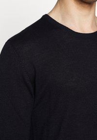 Czarny sweter z okrągłym dekoltem, na którym widać zbliżenie teksturowanej dzianiny, z gładką linią szyi i detalem szwu na ramieniu.