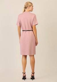 Rosa Strickkleid mit kurzen Ärmeln, knielang geschnitten, in der Taille mit einem schwarzen Gürtel tailliert. Kombiniert mit schwarzen Absatzsandalen.