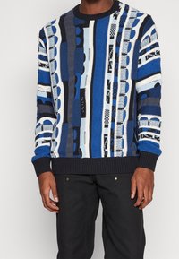 Gestickter Pullover mit auffälligen blauen, schwarzen und weißen vertikalen Streifen. Strukturiertes Muster und gerippte Bündchen und Saum mit Akzenten von Text.