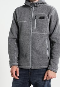 Szara polarowa kurtka z zamkiem błyskawicznym z przodu, kapturem i kontrastowymi szwami. Posiada dwie kieszenie z przodu oraz kieszeń na piersi z logo.