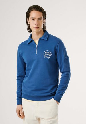 Modra zip-up mikica s ovratnikom, dolgimi rokavi in logotipom na prsih. Ima rebrast rob na rokavih in spodnjem delu, mehek material ter stranski žep.