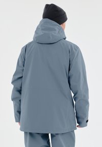 Grå vandtæt jakke med hætte, der har en glat tekstur, lange ærmer og en afslappet pasform. Minimalistisk design uden synlige logoer.
