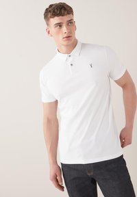 Valkoinen polo-paita, joka on valmistettu puuvillasta ja siinä on kauluspainike, kolme nappia, ja logo vasemmassa rinnassa. Slim fit -mallinen, lyhyt hiha.