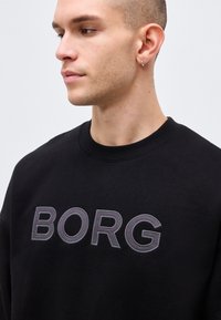 Νέος άνδρας με κοντά μαλλιά φοράει μαύρο φούτερ με μεγάλο γκρι κείμενο "BORG", κοιτάζει προς τα αριστερά σε απλό ανοιχτό γκρι φόντο.