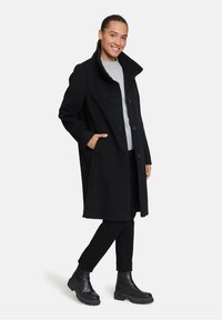 Cappotto lungo in lana nera con colletto alto, dotato di due tasche frontali e bottoni, abbinato a pantaloni neri e stivaletti alla caviglia.