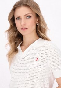 Biała koszulka polo z krótkim rękawem, z perforowaną teksturą i czerwonym haftem kotwicy na lewej piersi. Posiada klasyczny kołnierzyk i luźny krój.