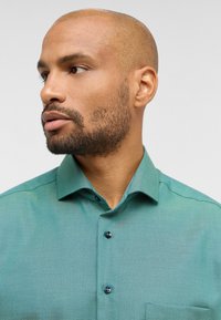 Camicia di cotone teal con colletto appuntito, chiusura con bottoni e texture sottile; presenta una fodera del colletto interno a contrasto e bottoni scuri.