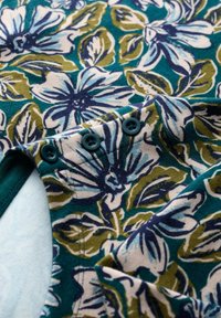 Materiał w kwiatowy wzór w odcieniach niebieskiego, zielonego i kremowego. Zawiera trzy turkusowe guziki na dekolcie oraz miękką strukturę.