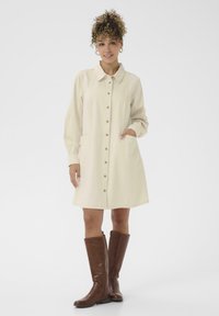 Krém színű ingruha gallérral, elöl gombos záródással, hosszú ujjakkal és oldalsó zsebekkel. Barna térd fölé érő csizmával párosítva.