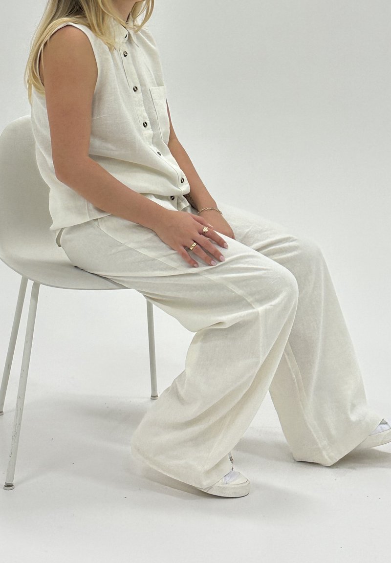 Vit linne ärmlös topp med knappstängning och ficka, kombinerad med vida vita linnebyxor. Accessoarer inkluderar flera ringar.
