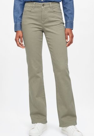 Person trägt beige gerade geschnittene Hose, blaues Denimhemd und weiße Sneaker, steht vor einem schlichten weißen Hintergrund.