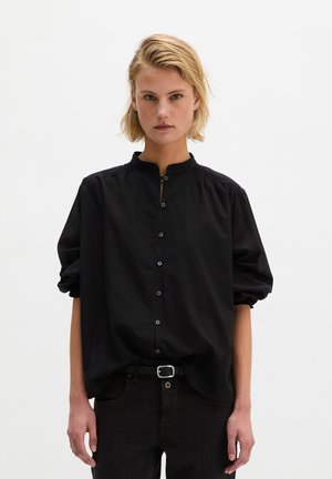 Femme aux cheveux blonds courts portant un chemisier noir boutonné avec des manches bouffantes et un pantalon noir, debout devant un fond blanc uni.