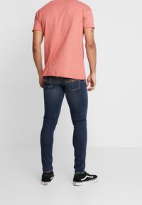 Herr T-shirt i rosa med kort ärm, vy bakifrån, ihopparad med mörkblå tajta jeans och svarta sneakers. Enkelt utseende, inga mönster.