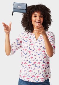 Witte blouse met een roze en blauwe bloemenprint, vier knopen bij de halslijn en driekwartmouwen; blauwe portemonnee zwevend erboven.