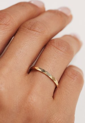 Gros plan sur une main portant une fine bague en or facettée à l'annulaire, contre un fond neutre.