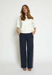 White cropped sweatshirt with a ribbed hem, paired with navy wide-leg pants featuring vertical stripes. Model standing, hands in pockets.