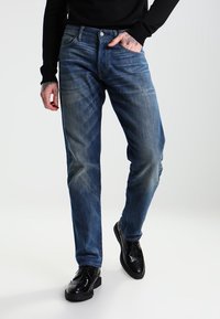 Vaqueros de mezclilla azul oscuro con corte recto, que presentan un ligero desvanecimiento, cinco bolsillos y un cierre de botones. Combinados con zapatos de vestir negros.