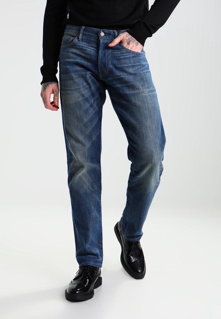 Vaqueros de mezclilla azul oscuro con corte recto, que presentan un ligero desvanecimiento, cinco bolsillos y un cierre de botones. Combinados con zapatos de vestir negros.