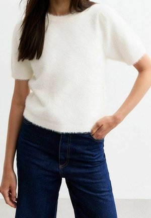 Nainen pukeutuneena valkoiseen pörröiseen lyhythihaiseen neulepaitaan ja tummansinisiin korkeavyötäröisiin farkkuihin, toinen käsi taskussa.