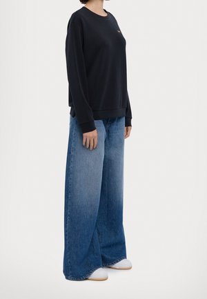 Person wearing black long-sleeve sweatshirt, wide-leg blue jeans, and white sneakers, standing against a plain light background.