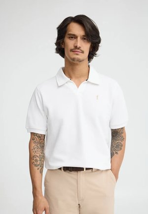 Mężczyzna o średniej długości ciemnych włosach i tatuażach na ramionach, ubrany w białą koszulkę polo i beżowe spodnie, stojący z jedną ręką w kieszeni.