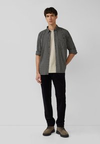 Herren kariertes Hemd in Schwarz und Weiß, zugeknöpft, mit hochgekrempelten Ärmeln; kombiniert mit einem hellbeigen T-Shirt und dunklen Hosen. Braune Schnürstiefel.