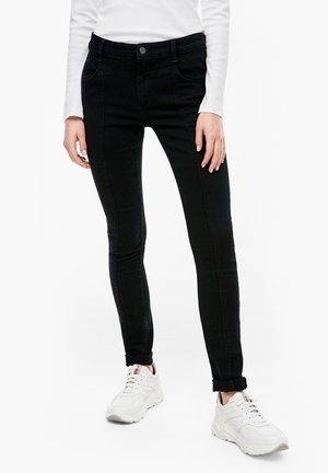 Jean skinny noir avec une texture lisse, comportant des poches avant et une fermeture à bouton, associés à des chaussures de sport blanches.