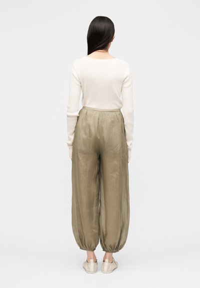 Femme aux longs cheveux foncés tournée vers l’arrière, portant un haut blanc à manches longues, un pantalon ample kaki avec poignets froncés et des chaussures claires.