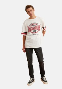 Camiseta blanca de manga corta con un gráfico de varsity rojo y acentos de franjas negras. Combinada con unos jeans oscuros y zapatillas negras.