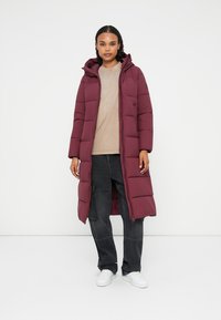 Caban lung bordeaux cu umplutură, cu glugă, având secțiuni matlasate, închidere cu fermoar și buzunare laterale, purtat peste un top bej și pantaloni negri.