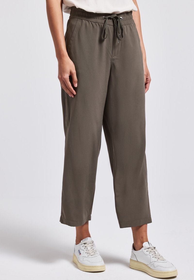 Personne portant un pantalon ample vert olive à cordon et des baskets blanches, debout devant un fond clair uni.