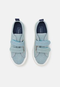 Baskets en denim bleu clair avec deux bandes velcro, semelles en caoutchouc blanc et étiquette de semelle intérieure bleu marine. Forme arrondie avec des surpiqûres accentuées.