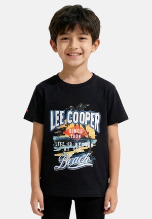 Garçon souriant aux cheveux foncés portant un t-shirt noir Lee Cooper avec un coucher de soleil, un palmier et le texte "La vie est meilleure à la plage."