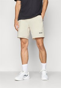 Pantalones cortos deportivos beige con una cintura elástica, que cuentan con bolsillos laterales y un logo negro "BOSS" en el muslo izquierdo. Tejido suave y ligero.
