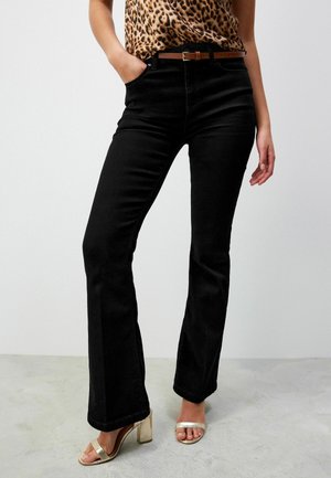 Femme portant un pantalon flare noir taille haute, ceinture marron, haut imprimé léopard et sandales dorées à talons blocs ouverts, debout sur un sol en béton.