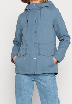 Femme portant une veste à capuche bleue avec des boutons-pression sur le devant, une poche zippée, des cordons, des poches à rabat et un jean bleu clair, les mains dans les poches de la veste.