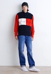 Langärmliges Polohemd in Marineblau, Rot und Weiß mit einem Farbblockdesign und Logo. Kombiniert mit blauen, ausgewaschenen Jeans und weißen Sneakers, ergänzt durch eine schwarze Mütze.