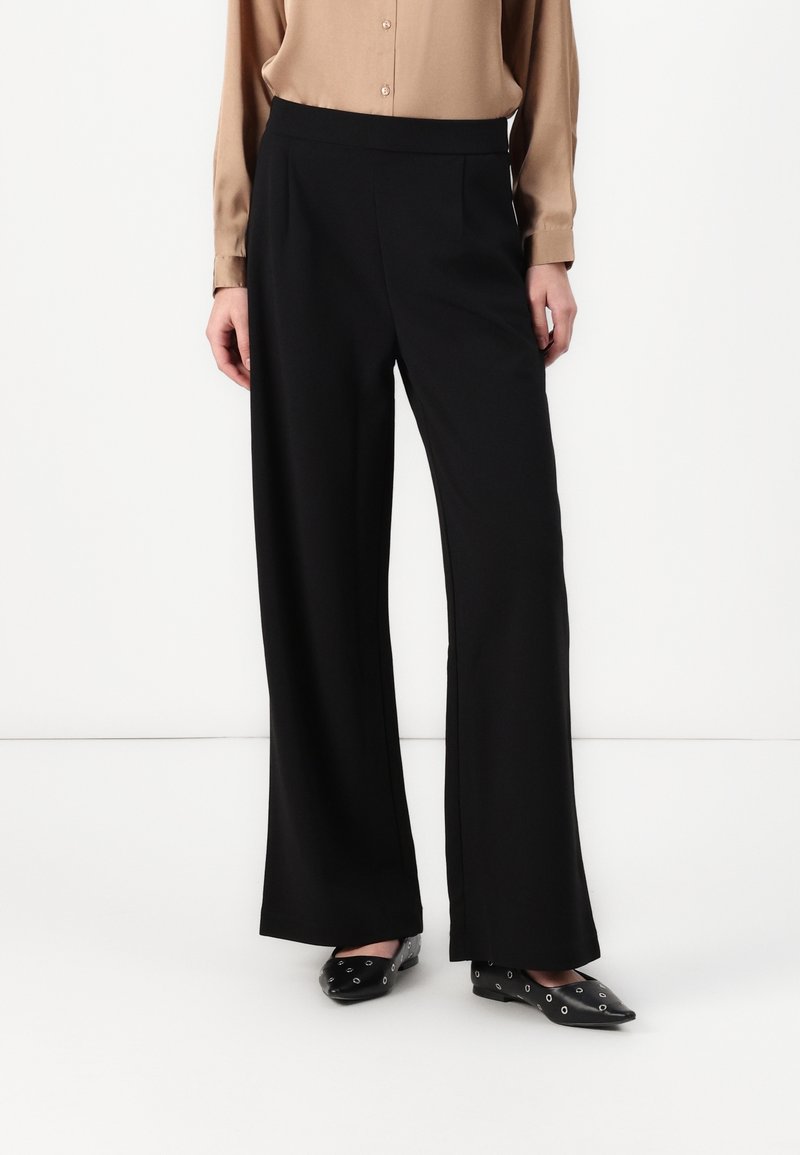 Personne portant un pantalon noir à jambes larges, un chemisier beige à manches longues et des chaussures noires pointues avec des œillets argentés.
