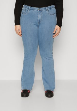 Persona que lleva jeans vaqueros de mezclilla azul claro combinados con zapatillas negras con rayas rojas, de pie frente a un fondo claro y sencillo.