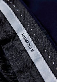 Lindbergh-Markenlabel auf einem dunklen Stoff mit einem strukturierten schwarzen Hintergrund und einem silbernen Streifenakzent, verziert mit weißen Nähdetails.