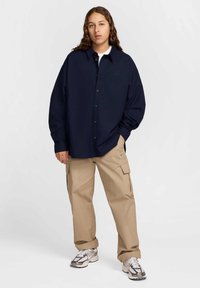 Camisa azul-marinho oversized com botões e mangas longas, combinada com calças cargo bege com bolsos. Ténis atléticos cinzentos nos pés.