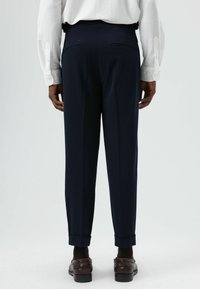 Pantalon marine à coupe ajustée, présentant une texture lisse, des plis et des ourlets retroussés. Poches arrière visibles, porté avec des chaussures marron.